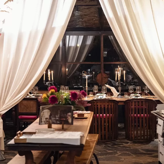 A cosy, elegantly set dining table in a dimly lit restaurant, framed by flowing white curtains. The wooden table is adorned with candles, glassware, and floral centerpieces, creating an inviting and intimate atmosphere.