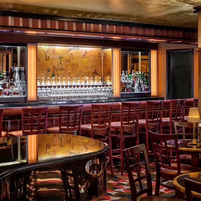 A cozy, dimly lit bar with a long counter and a row of barstools. Behind the counter are shelves stocked with various bottles and glasses. The ceiling is mirrored, and the floor has a colorful patterned carpet.