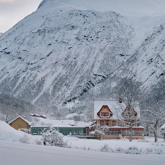 A picturesque winter scene with a large, red wooden house surrounded by snow-covered trees and buildings. In the background, a vast mountain rises, blanketed in snow under a cloudy sky.