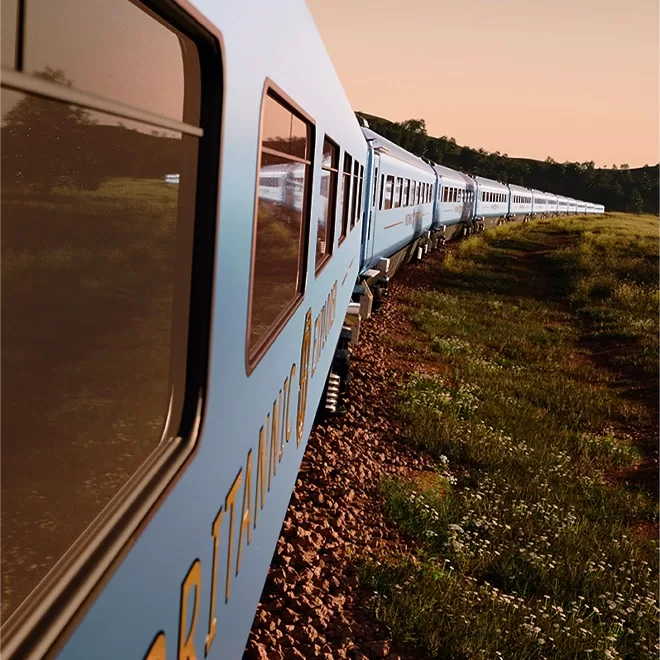 A long, blue train winds through a lush green landscape under a clear sky. The sun casts a warm glow over the scene, highlighting the serene countryside and the train’s reflective windows as it curves along the tracks.