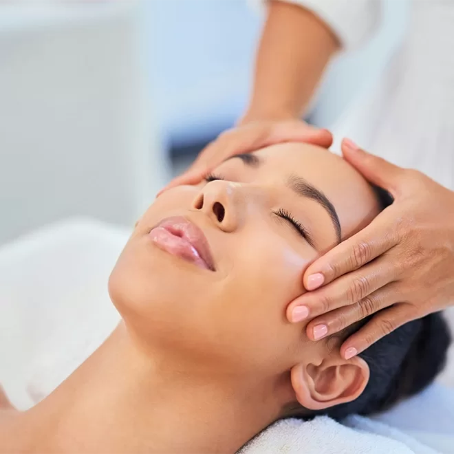 A person receiving a facial massage, with eyes closed, lying on a spa table. Their head is gently held by the masseurs hands, creating a relaxed and serene atmosphere.