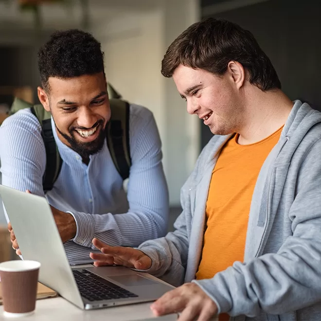 Two men smile at a laptop screen. The one on the left wears a backpack and light blue shirt. The one on the right, wearing a gray hoodie over an orange shirt, points at the screen. A paper cup is on the table.