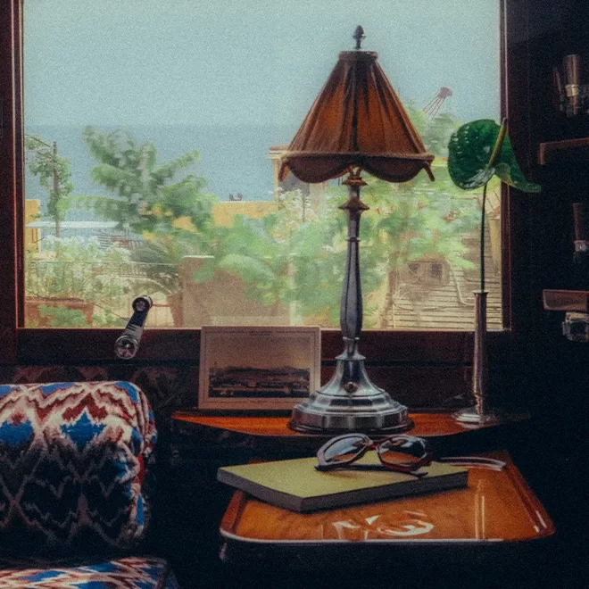 Cosy room aboard the Venice Simplon-Orient-Express, a train by Belmond, with a vintage lamp on a wooden table beside a patterned chair. Sunglasses and a notebook sit on the table. A window reveals a lush garden with plants outside, creating a peaceful atmosphere. A framed photo and various decor items complete the scene.