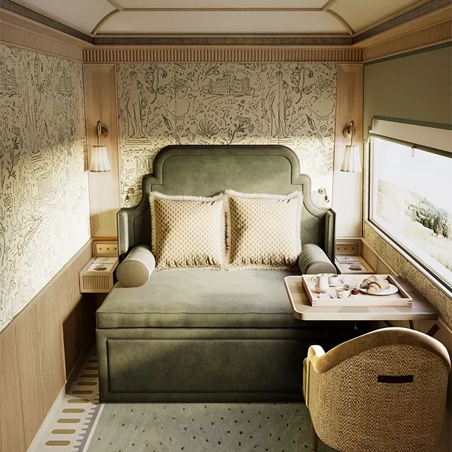 Luxurious train cabin inside the Britannic Explorer, a train by Belmond, with a plush green bed, matching padded headboard, and two beige pillows. The room features light wood paneling, decorative wallpaper, a window with a scenic view, and a wooden tray on the bed. A cushioned chair is nearby.
