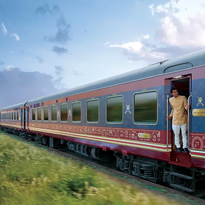 The luxury train - the Deccan Odyssey in India, adorned with intricate designs travels through lush greenery under a partly cloudy sky. A person stands at the entrance of a carriage, looking outside as the train moves.