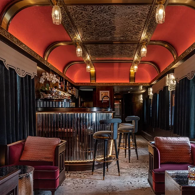 Elegant vintage-style bar inside Silk Road Express train compartment by Train of Glamour in China, with velvet armchairs, high stools, and a curved counter. The ceiling features intricate designs and warm lighting from pendant lights. Dark drapes line the walls, adding to the luxurious ambiance.