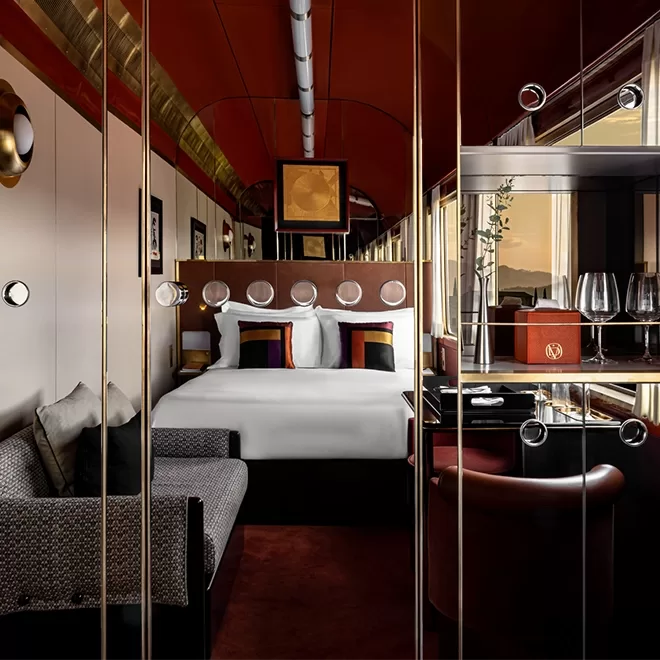 Luxurious train of La Dolce Vita Orient Express in Italy cabin interior featuring a neatly made bed with plush pillows, elegant decor with modern accents, a cosy sofa, and a stylish tray with glasses on a small table. Warm lighting enhances the sophisticated ambiance.