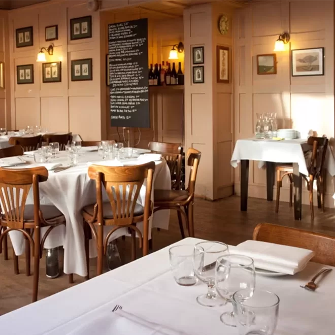 Cosy restaurant interior with wooden chairs and tables covered in white tablecloths. Framed art and dim wall lights adorn the paneled walls. A blackboard with menu items is featured, and wine bottles are displayed on a shelf at Bouchon Racine, Farringdon.