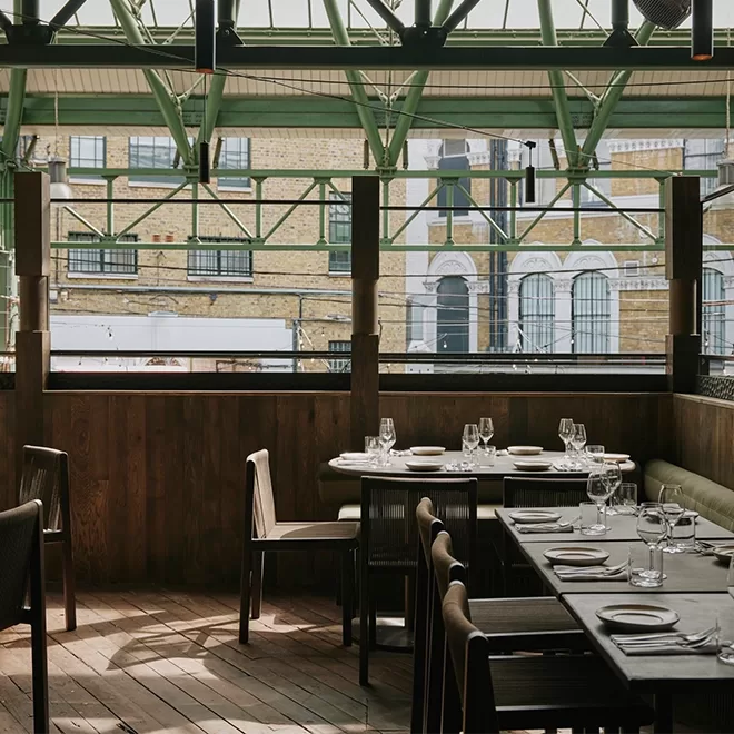 A modern restaurant interior featuring wooden flooring and tables set with glassware and napkins. Large windows reveal an urban brick building outside. The ceiling is supported by green metal beams. Tables and chairs are arranged neatly at OMA, Borough Market.