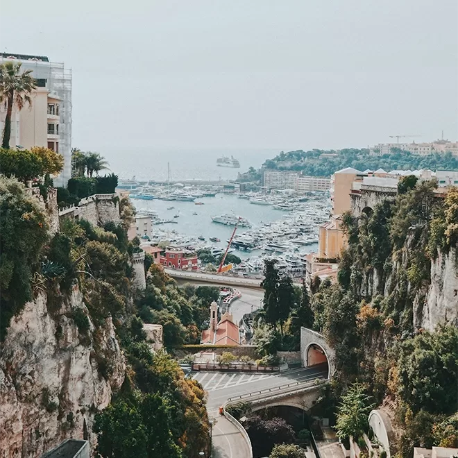 A scenic view of a harbour in Monaco with numerous boats and yachts, surrounded by buildings and lush greenery. A road winds through the rocky landscape, leading towards the waterfront. The sky is overcast, and the distant horizon is hazy.