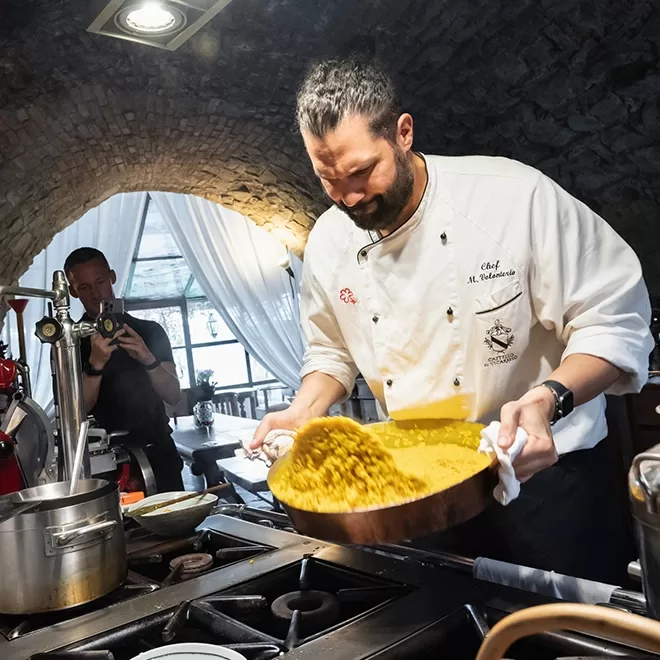 Head chef Massimiliano Volonterio, preparing a large pan of risotto in a rustic kitchen, under a stone archway. A man in the background takes a photo, capturing the lively culinary scene.