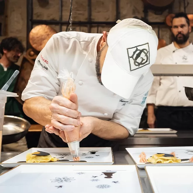 Chef, Francesco Bracali in a white uniform and cap carefully pipes sauce onto a plate in a busy kitchen, with other chefs in the background preparing dishes. The focus is on the detailed presentation of the food.