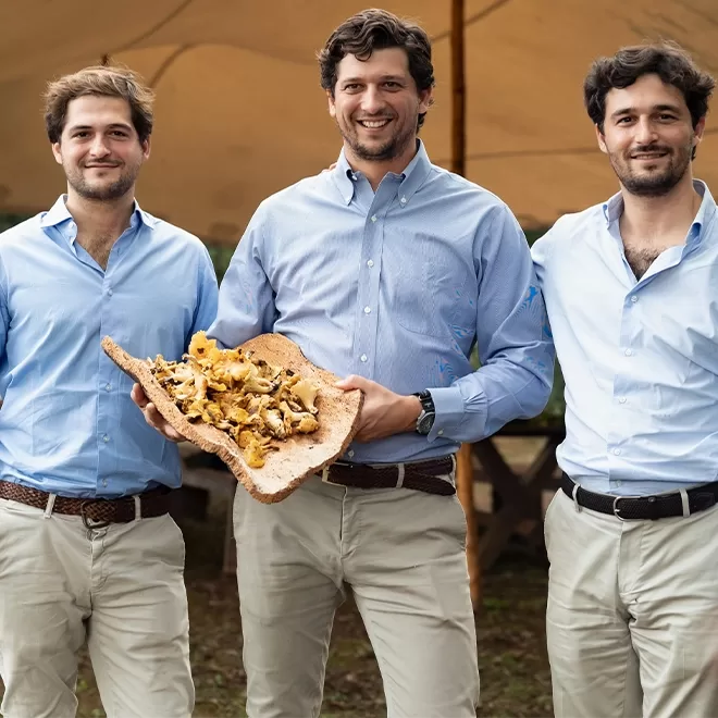 Three men - Corso, Brando, and Neri in blue shirts and beige pants stand outdoors under a canopy. The man in the center is holding a large wooden platter with a variety of mushrooms. They are smiling and appear to be at a gathering or event.