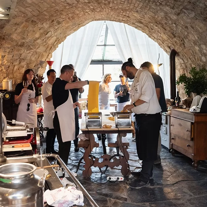A group of people in a rustic kitchen with stone walls are gathered around a table making pasta. Some are wearing aprons, and a chef is guiding them. Sunlight streams through a large window with white curtains in the background.