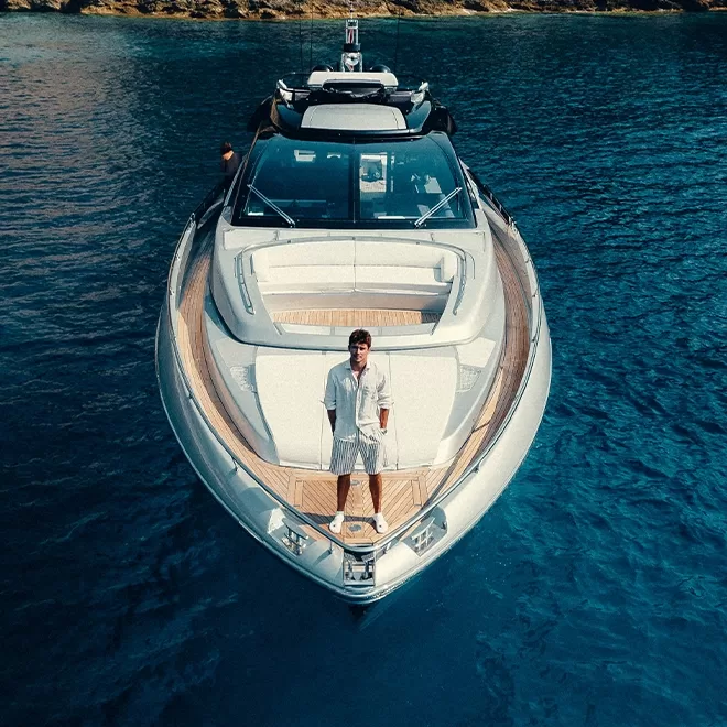 Charles Leclerc stands at the bow of a luxury yacht floating on clear blue water. The yacht is sleek and modern, featuring a light wood deck and white accents. The background shows a rocky shoreline under a clear sky.