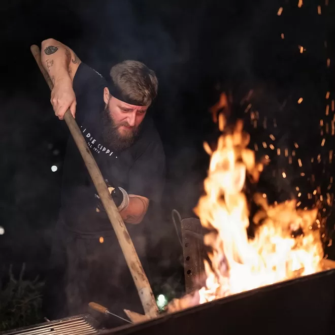 A bearded man wearing a headband and black shirt tends to a large outdoor grill at night, using a long-handled tool to adjust wood or charcoal, with flames rising dramatically in front of him.