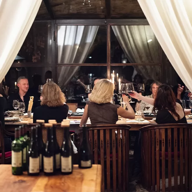 A group of people sitting around a dining table in a cosy, dimly-lit room with large windows and white curtains. Candles and glasses of wine are on the table, creating a warm and inviting atmosphere. Bottles are on a wooden surface in the foreground.