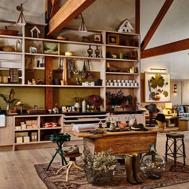A cosy rustic store within Wildflower Farms features wooden shelves filled with various home goods, decorations, and ceramics. A wooden table displays more items like a vase and boots. The warm interior is enhanced by wooden beams and earthy tones, echoing the charm of the Auberge Resorts Collection, New York.