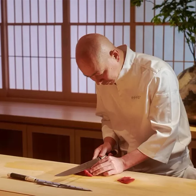 Chef in a white uniform skillfully slices fish on a wooden counter in a traditional Japanese setting, with a bonsai and shoji screen in the background. A long knife rests on the counter beside him at Sushi Kanesaka, London.