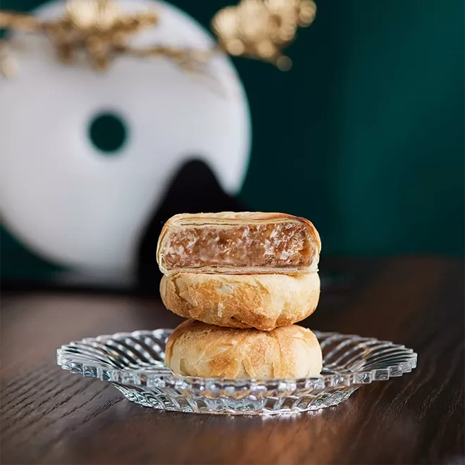A glass plate featuring three mooncakes, each adorned with intricate details and vibrant hues by The Merchants, Hong Kong.