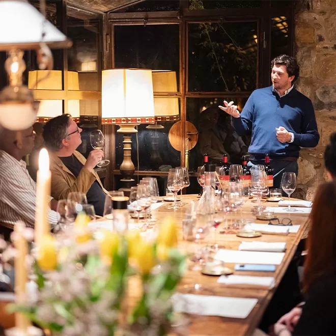 A man gestures while speaking to a group seated around a long wooden table set with wine glasses, bottles, and papers. The room has warm lighting, a stone wall, and large windows. Candles and flowers add to the cosy atmosphere.