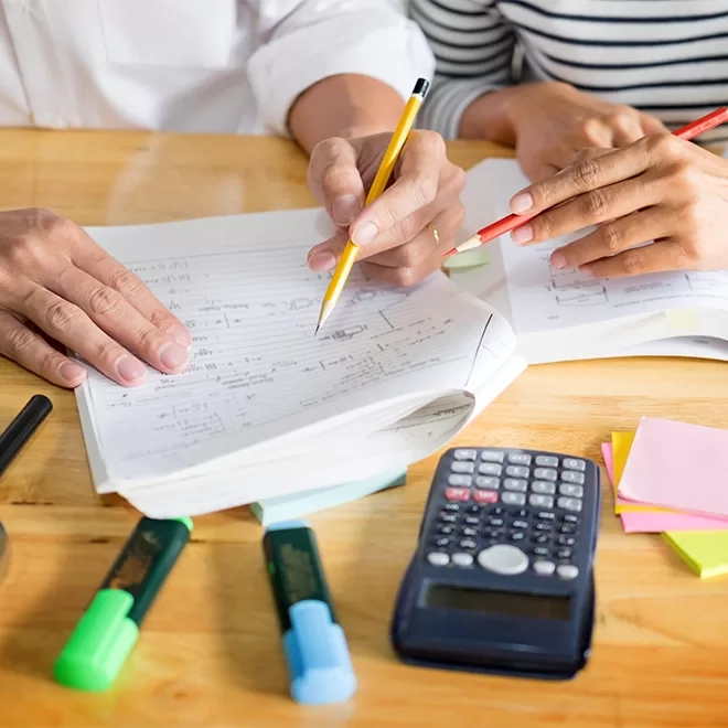 Two people sit at a table with open notebooks, pencils, and highlighters. A calculator and sticky notes are nearby. They appear to be collaborating on a project or studying together.
