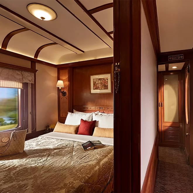 Luxurious train cabin of Deccan Odyssey inIndia, with a cosy bed, wooden paneling, and elegant decor. A bag sits on the bed, and a framed picture hangs on the wall. Soft lighting enhances the warm ambiance, and a window shows a blurred view of scenery passing by.