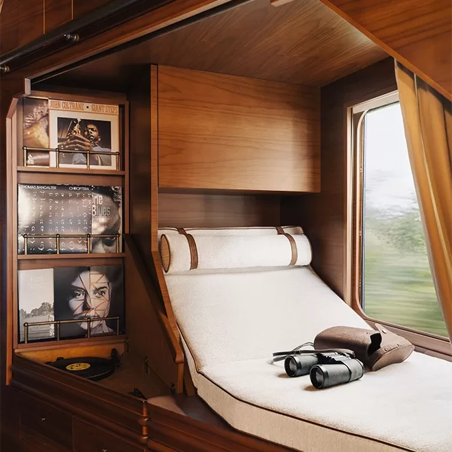 The cosy train cabin of Venice Simplon-Orient-Express, A Belmond Train in Europe with, a wooden interior features a plush reclining seat. On the wall are shelves holding vinyl records. A pair of binoculars is placed on a small table near the window, with a view of blurred outdoor scenery.