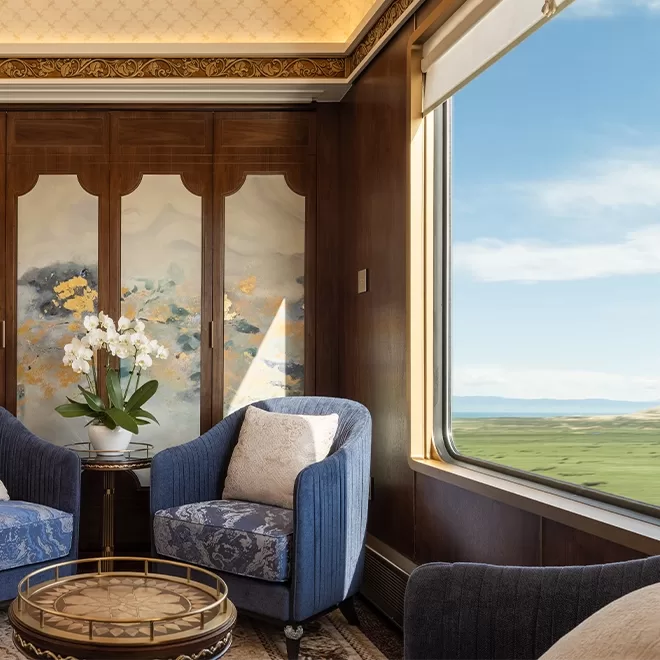 Luxurious Silk Road Express train compartment by Train of Glamour in China with elegant decor. Features two blue armchairs, a round glass table, and a decorative panel with floral art. A large window offers a scenic view of green fields and a clear blue sky. White orchids on the table.
