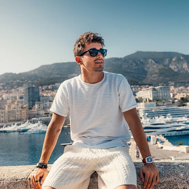 Charles Leclerc wearing sunglasses and a white shirt sits on a ledge overlooking a scenic harbor with boats. Hills and buildings are visible in the background under a clear blue sky.