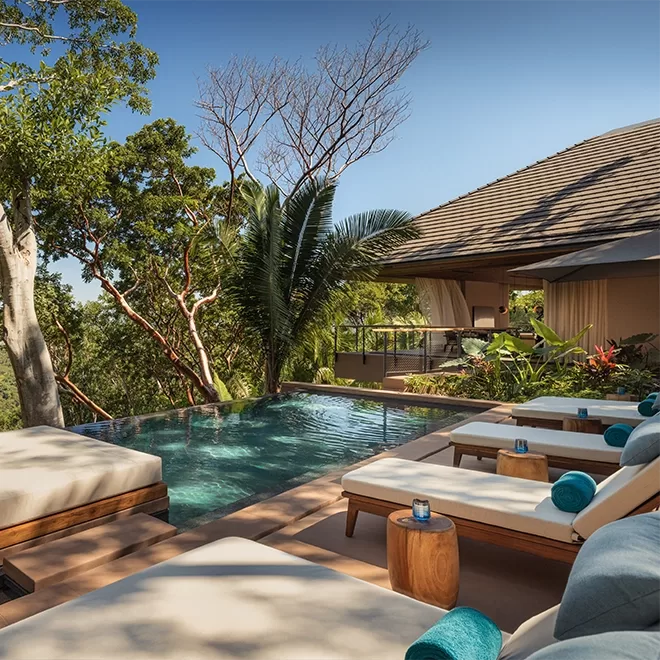 Luxurious outdoor space featuring a row of lounge chairs with blue cushions beside an infinity pool. Lush greenery surrounds the area, and a modern building with a sloped roof is visible in the background under a clear blue sky.