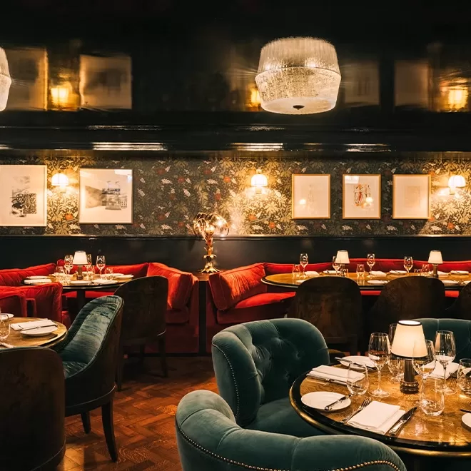 Elegant restaurant interior with plush red and green velvet chairs, round tables set with glassware and plates, floral wallpaper, framed art on the walls, and soft ambient lighting from wall sconces and chandeliers.