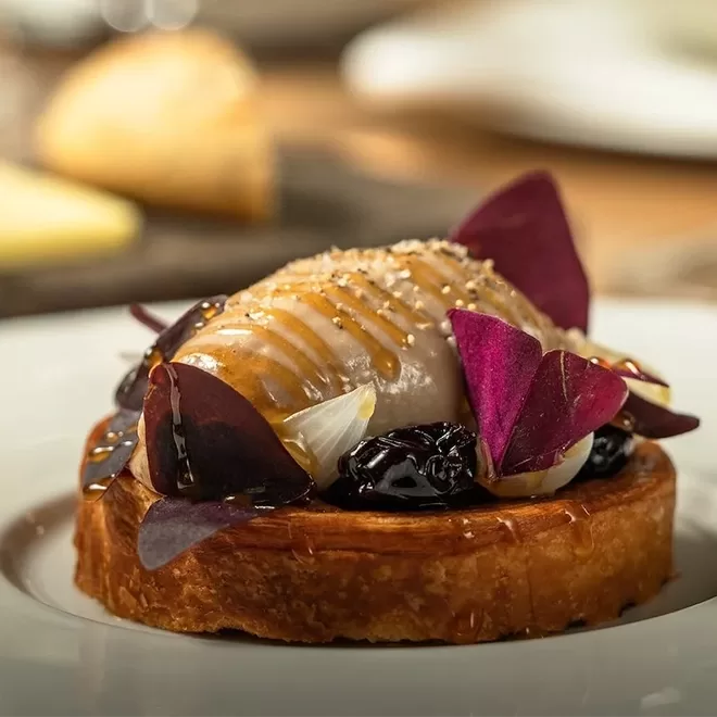 A gourmet tart topped with a swirl of honey, dark cherries, edible purple leaves, and a glossy glaze, presented on a white plate.