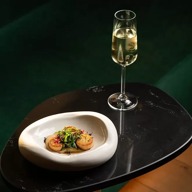 A white bowl with seared scallops and microgreens sits on a black marble table next to a tall glass of sparkling white wine against a dark green background.