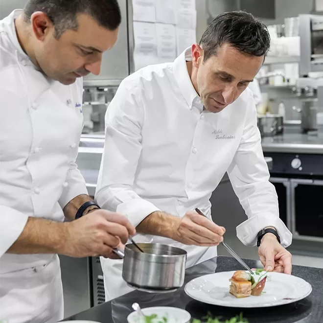 Two chefs in white jackets carefully plating a gourmet dish in a professional kitchen, one using tweezers for garnish while the other pours sauce from a small pot.