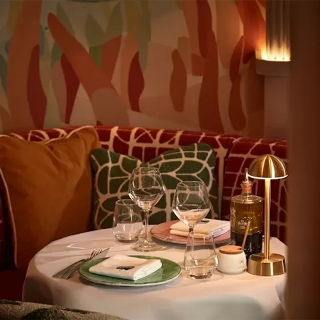 A cozy restaurant table for two with wine glasses, plates, and a small gold lamp, set against colorful pillows and a patterned wall.