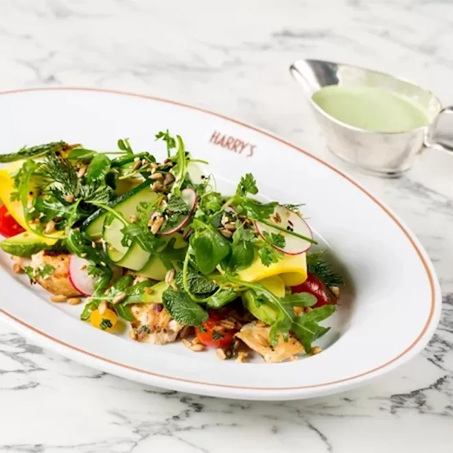 A fresh salad with grilled chicken, avocado slices, radishes, herbs, and greens is served on a white plate labeled HARRYS, with a small metal sauce boat of green dressing on the side.