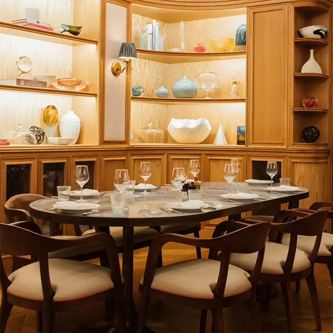 A stylish dining area with a set table for eight, surrounded by wooden chairs. Shelves in the background display decorative vases and art pieces, creating a warm, inviting atmosphere.