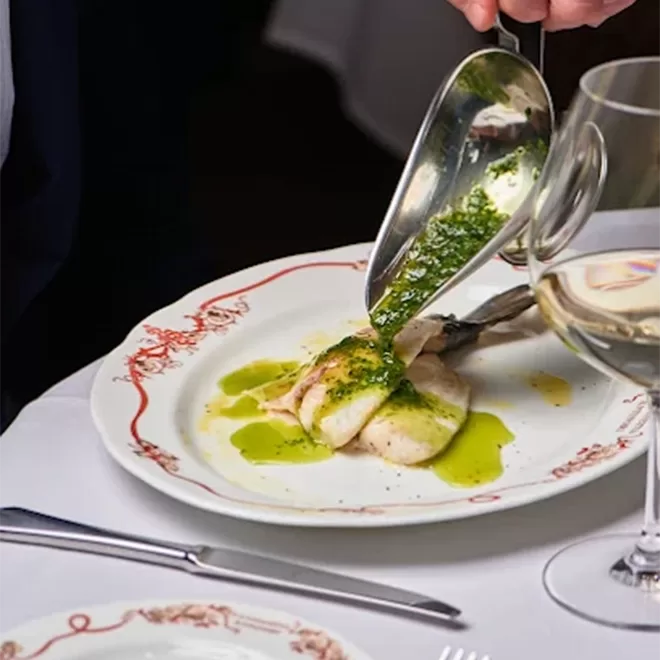 A hand pours green herb sauce over a plated fish fillet on a white dish with red detailing, beside a glass of white wine on a white tablecloth.