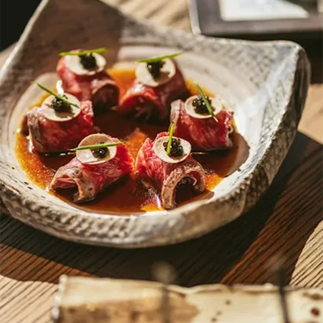 Five pieces of thinly sliced beef wrapped around a filling, topped with sauce, herbs, and a garnish, are arranged on a ceramic plate with brown sauce, set on a wooden table.