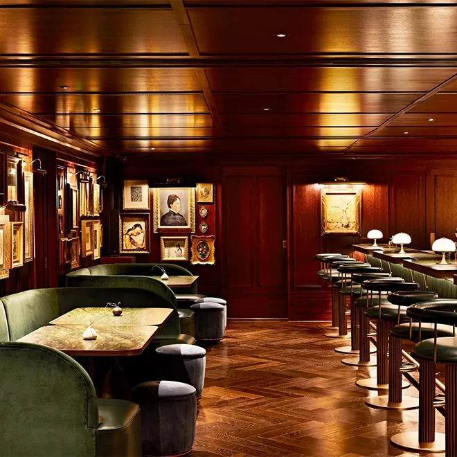 A sophisticated bar with dark wood paneling, green velvet booths, marble tables, barstools, and softly lit table lamps. Framed artwork decorates the walls, creating a warm, elegant atmosphere.