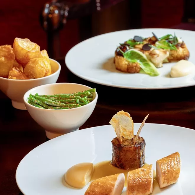 Plated gourmet meal with roast chicken, creamy sauce, and a decorative vegetable garnish, served alongside bowls of crispy potatoes and green beans; another elegant dish is visible in the background.