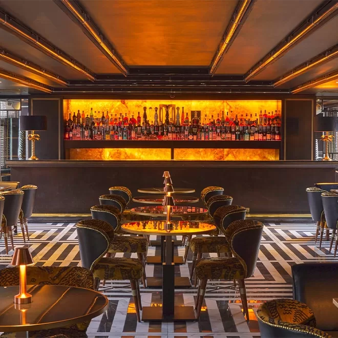 A stylish, modern bar with a glowing amber backlit shelf filled with bottles, elegant dark chairs around small tables, patterned floor, and warm ambient lighting.