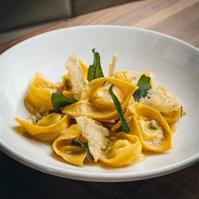 A white bowl filled with yellow tortellini pasta, garnished with crispy sage leaves and thin Parmesan crisps, sits on a wooden table.