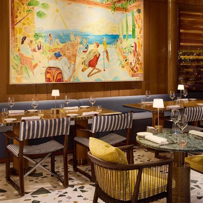 A stylish restaurant with striped chairs, set tables, and a large colorful painting depicting a lively beach scene on the wall above a blue cushioned bench. Warm lighting adds a cozy, inviting atmosphere.