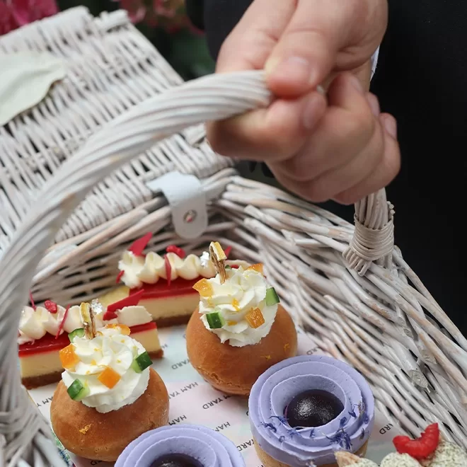 A hand holds a white wicker basket filled with assorted pastries, including purple tarts, cream-topped buns with fruit, and a rectangular layered cake with cream and berries.
