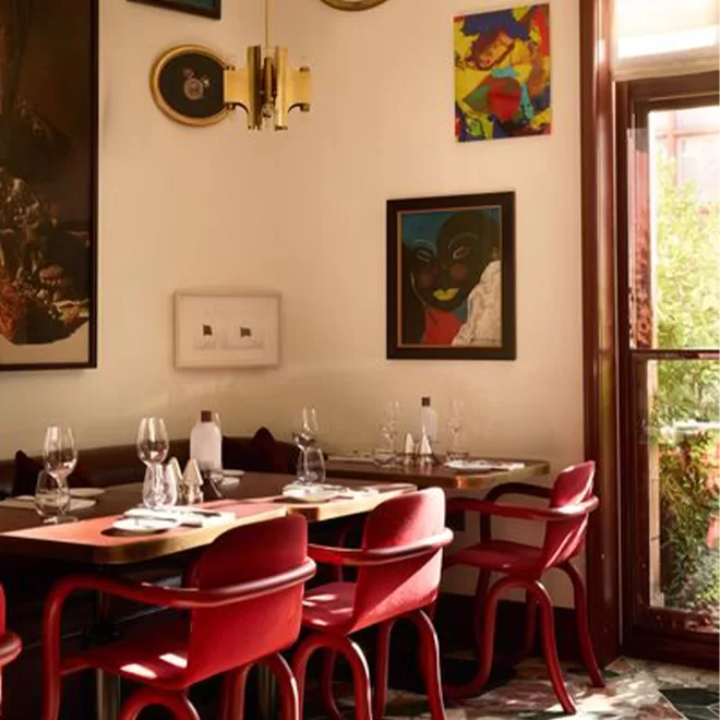 A cozy restaurant corner with red chairs, a wooden table set for four, colorful artwork on cream walls, and a large window letting in natural light. Wine glasses and tableware are neatly arranged.