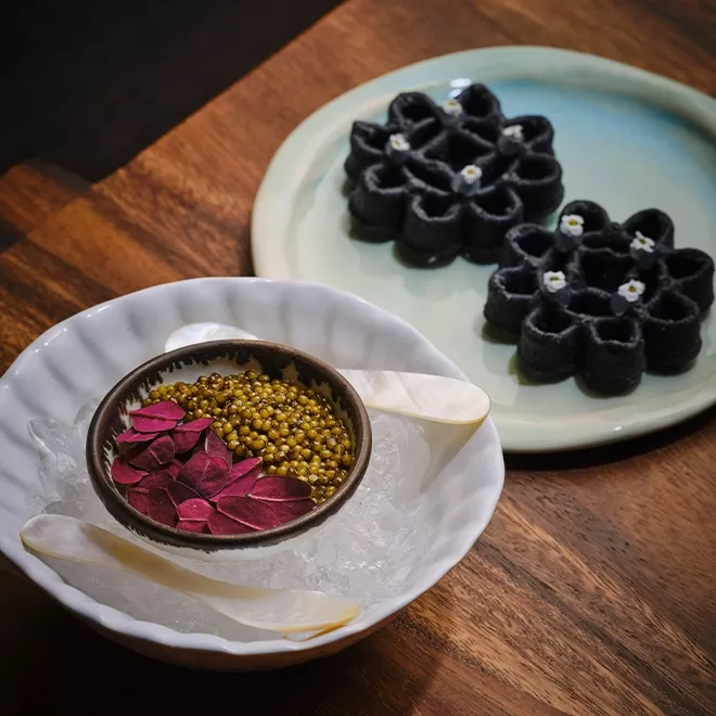 A bowl with golden caviar, red leaves, and two white spoons on ice, next to a plate with two black flower-shaped waffles topped with tiny white flowers, all set on a wooden table.
