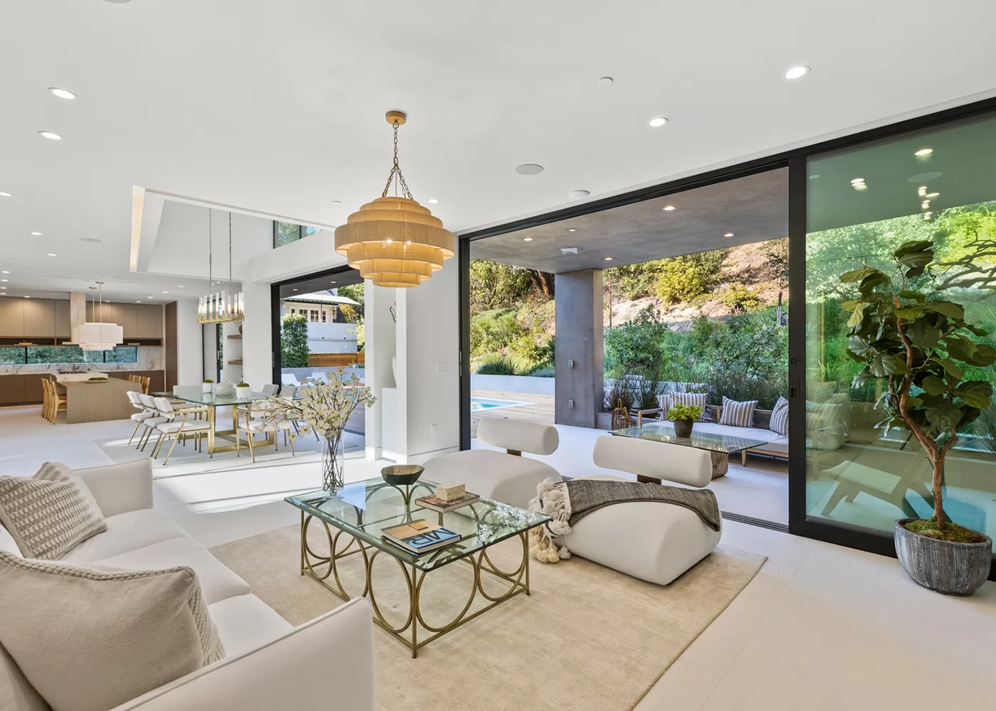 Modern open-concept living room with white furniture, glass coffee table, and gold chandelier. Large sliding doors open to a patio with seating, a pool, and lush greenery outside. Dining area and kitchen are visible in the background.