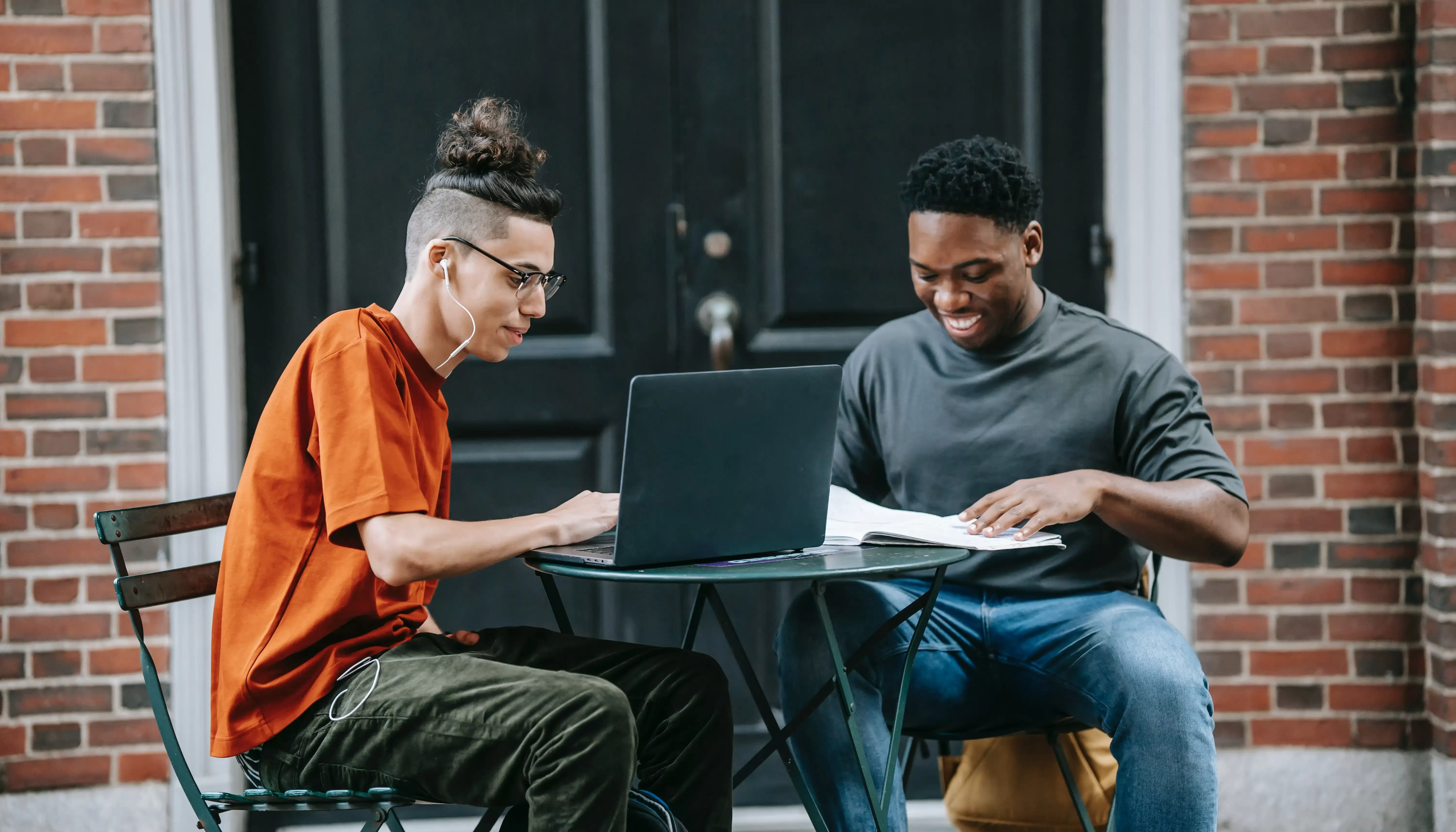 Student and tutor in casual clothing sat on an outdoor table in front of a building with a laptop and paper